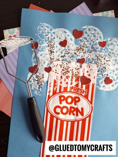 A popcorn-themed craft made with a blue paper base, red-and-white-striped paper bag, heart-shaped glitter, and white paint stamped to look like popcorn, with a potato masher beside it. Text reads @GLUEDTOMYCRAFTS.