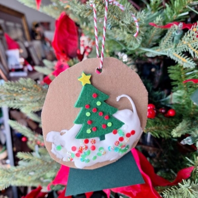 A handmade Christmas ornament hangs on a tree, featuring a green felt tree with colorful sequins, a yellow star, and a white cat at the base—reminiscent of puffy paint snow globe ornaments. Red and white twine secures it. @GLUEDTOMYCRAFTS is written below.