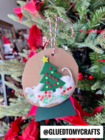 A handmade Christmas ornament with a felt tree, sequins, and a white cat design hangs on a decorated Christmas tree. Red poinsettias and greenery are in the background. The text @GLUEDTOMYCRAFTS is at the bottom.