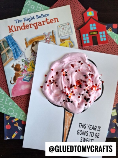 A children’s book titled The Night Before Kindergarten and a card with a puffy pink ice cream cone, decorated with heart-shaped sprinkles like puffy paint crafts, are surrounded by school-themed cutouts. The card says, This year is going to be sweet!.