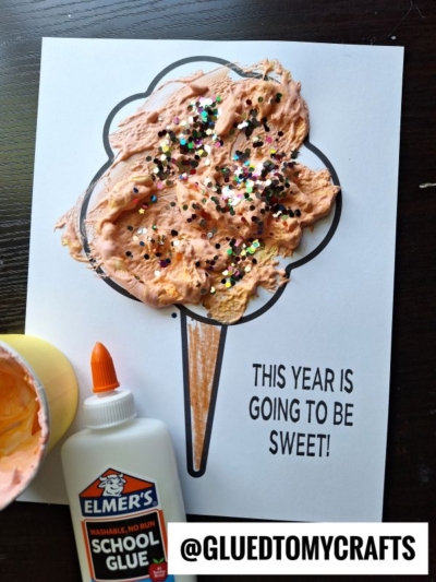 A puffy paint cotton candy craft made with an orange fluffy mixture and glitter on paper with THIS YEAR IS GOING TO BE SWEET! Glue bottle and cup with orange mixture are nearby. @GLUEDTOMYCRAFTS is at the bottom.