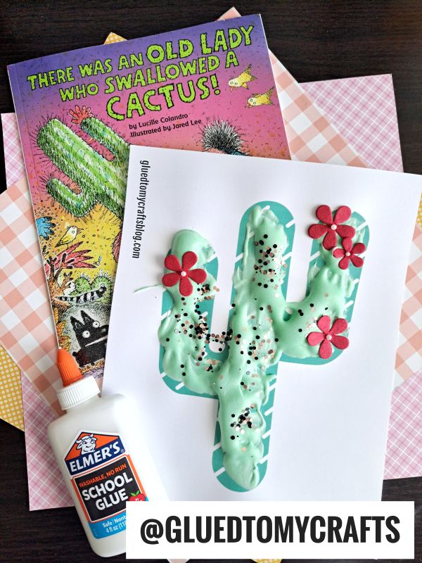 A children's book titled There Was an Old Lady Who Swallowed a Cactus! sits next to a puffy paint crafts cactus adorned with red flowers and black sprinkles. Elmer's glue and pastel papers are nearby.