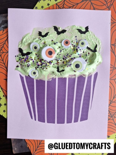 A Halloween-themed puffy paint witch cupcake craft with green puffy icing, googly eyes, bat shapes, and sprinkles, displayed on lilac paper with a purple cupcake liner illustration.