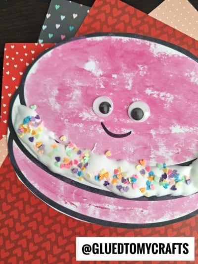 A pink, smiling puffy paint macaroon with googly eyes and colorful heart-shaped sprinkles sits on heart-patterned paper. The Instagram handle @GLUEDTOMYCRAFTS is at the bottom of the image.