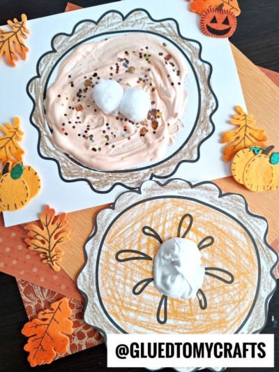 Two handmade puffy paint pie pumpkin crafts made with paper, orange-colored whipped topping, glitter, and cotton balls for whipped cream. Surrounded by felt pumpkins, leaves, and autumn-themed decorations. @GLUEDTOMYCRAFTS is written at the bottom.