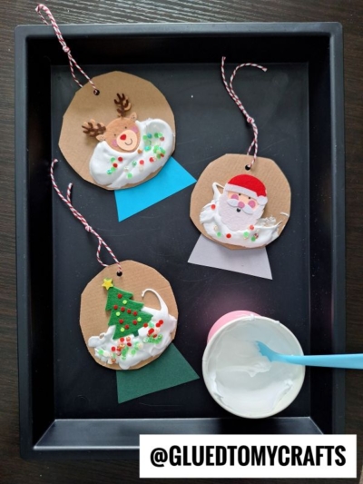 Three handmade Christmas ornaments with a reindeer, Santa, and a tree on brown circles, decorated with white glue and colorful beads, are displayed on a tray with a cup of white glue and a mixing stick. @GLUEDTOMYCRAFTS is labeled.