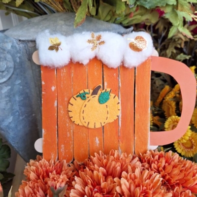 A handmade fall craft shaped like a mug, made of orange popsicle sticks with a pumpkin decoration, white “foam” on top, and surrounded by orange flowers. Text at the bottom reads @GLUEDTOMYCRAFTS.