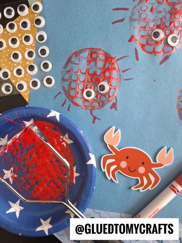 A blue paper with red paint stamps resembling crabs, made using a fly swatter. Googly eyes are added to the stamps, with a smiling cartoon crab, googly eyes sheet, and a marker nearby. @GLUEDTOMYCRAFTS is written at the bottom.