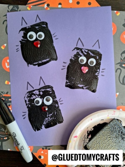 Purple paper features three black cats made from textured halloween spatula stamped crafts, each with googly eyes and a red nose. Black marker ears and whiskers are drawn on. A marker, a paint plate, and Halloween-themed paper are nearby.