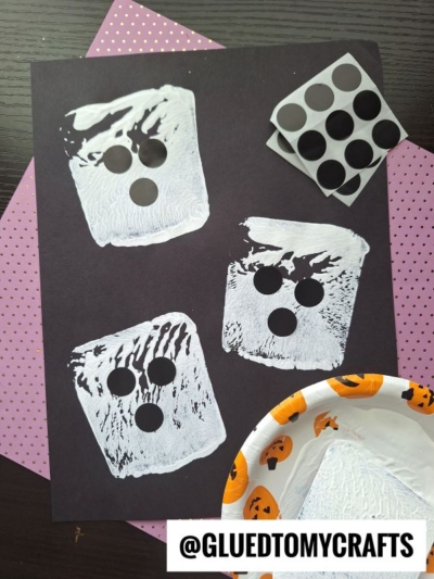 A black paper with three white ghost shapes, each with three black circle stickers for eyes and mouth. A fourth ghost shape is partly off the paper. Nearby are a polka-dot paper, sticker sheet, and Halloween plate.
