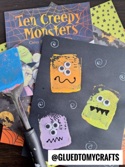 A Halloween-themed craft scene with a black paper featuring three colorful monsters with googly eyes, a spatula with blue paint, a Ten Creepy Monsters book, and patterned papers. @GLUEDTOMYCRAFTS is at the bottom.