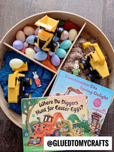 A round wooden tray holds colorful plastic eggs, toy construction vehicles, small animal figurines, blue and brown sensory sand, and two children’s books about diggers—a fun story time sensory bin idea for creative play.