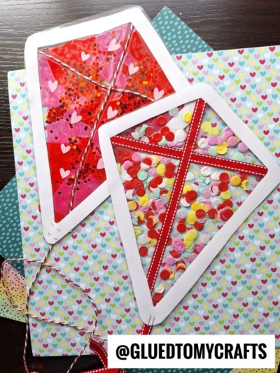Two stained glass kite designs, decorated with red, pink, and white confetti and heart shapes, sit on heart-patterned paper. Each paper kite has a white border, red ribbon accents, and twine tails. @GLUETOMYCRAFTS is labeled at the bottom.
