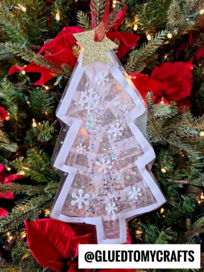 A handmade stained glass Christmas tree ornament with a white border, filled with shiny tissue pieces, snowflakes, and glitter, topped with a gold star, hangs on a decorated Christmas tree. @GLUEDTOMYCRAFTS is written below.