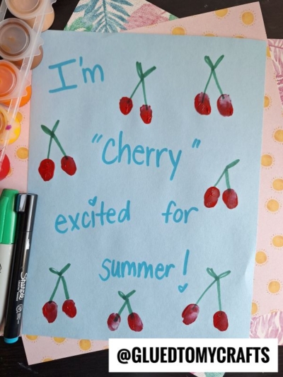 A blue paper with Im cherry excited for summer! written in blue marker. Red cherry drawings and craft supplies, including markers and paint, are around the paper—perfect for end of school year keepsakes. @GLUEDTOMYCRAFTS is at the bottom.