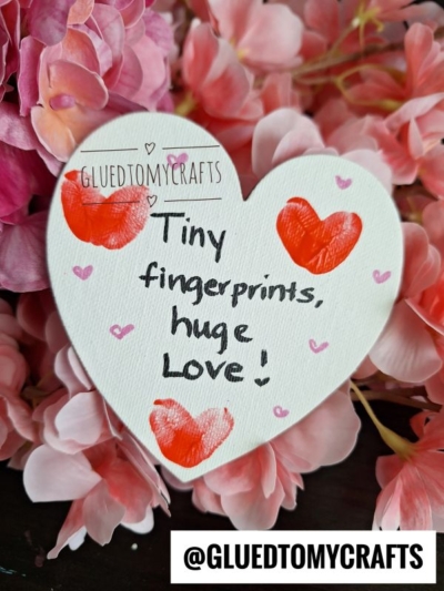 A white heart-shaped card with red fingerprint hearts and the handwritten words Tiny fingerprints, huge love! sits on pink flowers. Perfect for THUMBPRINT CRAFT IDEAS FOR SPRING, it's labeled GLUEDTOMYCRAFTS. Bottom text: @GLUEDTOMYCRAFTS.