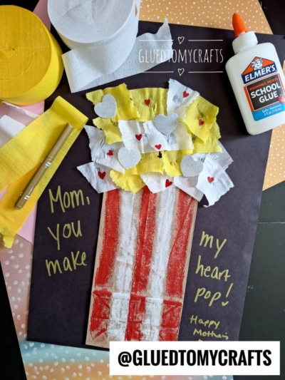 A handmade popcorn craft on black paper, with red and white stripes and yellow-and-white tissue popcorn, heart cutouts, glue, scissors, and a note reading, Mom, you make my heart pop! Happy Mother’s Day.
