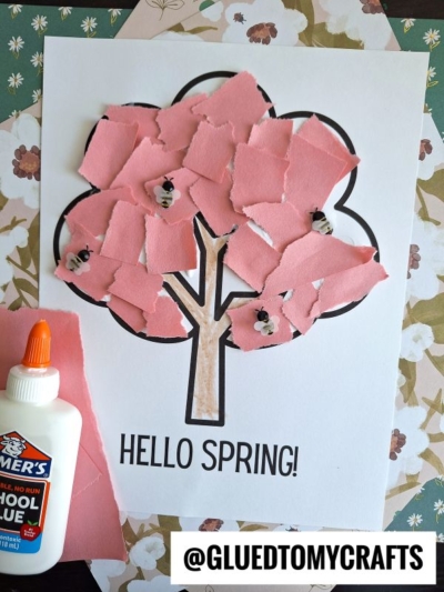 A spring craft project featuring a tree outline on paper with “HELLO SPRING!” written below. The tree’s canopy is made of pink torn paper pieces, with small bees drawn among them. A bottle of Elmer’s glue is nearby.