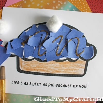 A handmade pie craft made with torn blue paper, cotton balls, and glitter glue on a printed template that reads LIFES AS SWEET AS PIE BECAUSE OF YOU! Supplies like glue and colored paper are nearby.