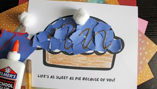 A handmade pie craft made with torn blue paper, cotton balls, and glitter glue on a printed template that reads LIFES AS SWEET AS PIE BECAUSE OF YOU! Supplies like glue and colored paper are nearby.