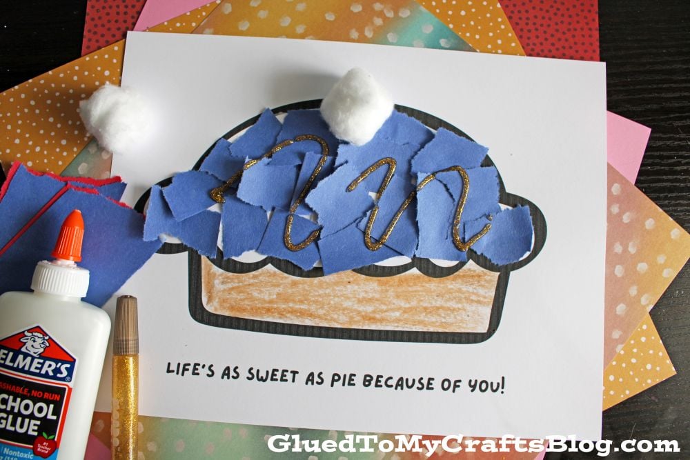 A handmade pie craft made with torn blue paper, cotton balls, and glitter glue on a printed template that reads LIFES AS SWEET AS PIE BECAUSE OF YOU! Supplies like glue and colored paper are nearby.