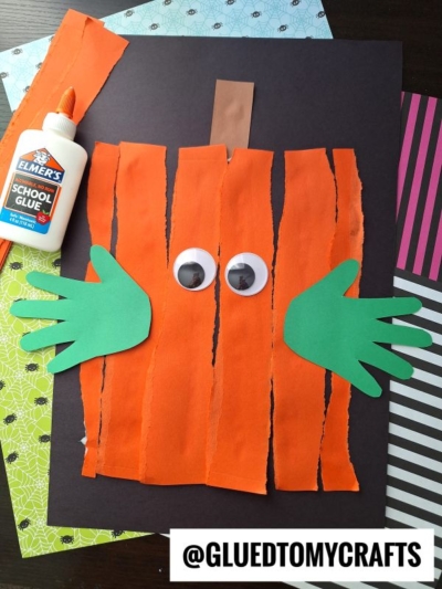 A pumpkin craft made from ripped orange paper strips, two green handprint cutouts for leaves, two large googly eyes, and a brown stem, all glued to black paper. A bottle of Elmer’s glue is shown beside the craft.