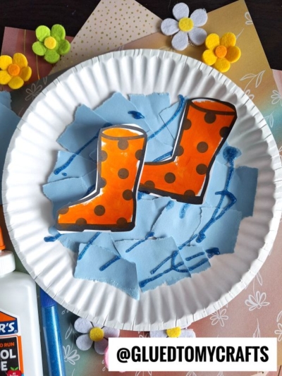 A paper plate features an art project with orange polka-dot rain boots made from paper, placed on blue torn paper pieces meant to look like water. Colorful paper flowers and craft supplies are nearby.