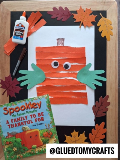 A fall-themed craft setup with a book titled “Spookley the Square Pumpkin,” a square pumpkin art made of orange paper strips, green handprint leaves, googly eyes, glue, marker, and artificial autumn leaves.