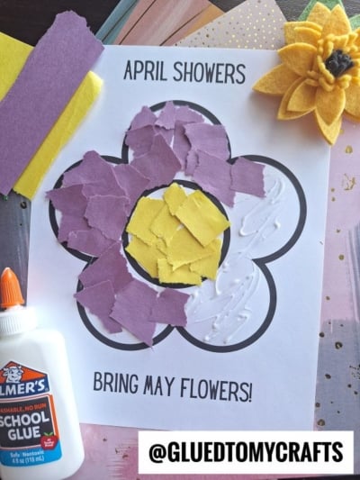 A children’s craft features a flower outline filled with torn purple and yellow paper. Text reads APRIL SHOWERS BRING MAY FLOWERS! Nearby are a glue bottle, felt flower, and colored paper. Username @GLUEDTOMYCRAFTS is shown.