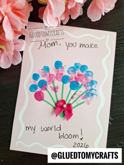 A handmade card on a dark surface features pink and blue flower art, a pink ribbon bow, and the message Mom, you make my world bloom! 2026. Perfect for THUMBPRINT CRAFT IDEAS FOR SPRING. Pink flowers and @GLUETOMYCRAFTS are visible nearby.