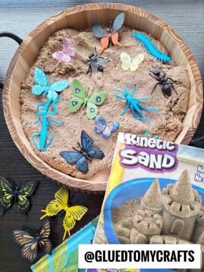 A round wooden tray filled with kinetic sand and various colorful plastic insects is shown. The Kinetic Sand package and a few plastic butterflies are placed next to the tray. @GLUETOMYCRAFTS is written at the bottom.