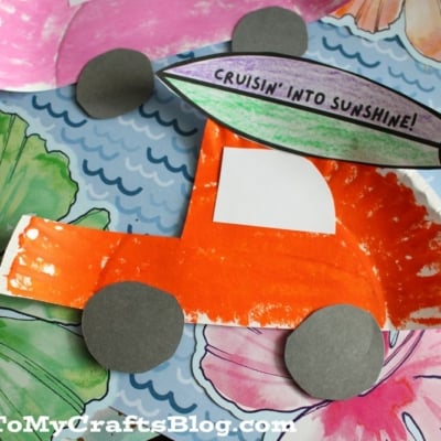 A colorful paper craft of an orange car with gray wheels made from a painted paper plate, decorated with a surfboard saying Cruisin into Sunshine! sits on a floral background.