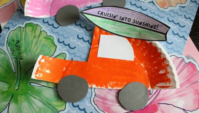 A colorful paper craft of an orange car with gray wheels made from a painted paper plate, decorated with a surfboard saying Cruisin into Sunshine! sits on a floral background.