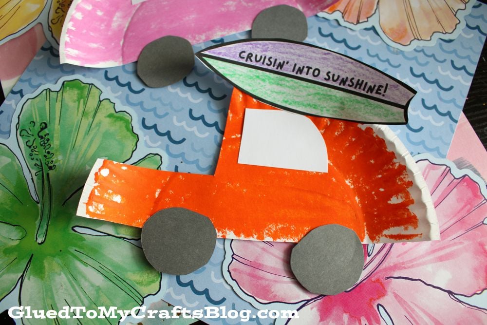 A colorful paper craft of an orange car with gray wheels made from a painted paper plate, decorated with a surfboard saying Cruisin into Sunshine! sits on a floral background.