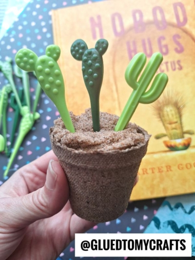 A hand holds a small brown pot filled with sand and three green cactus-shaped drink stirrers. A book titled “Nobody Hugs a Cactus” is in the background. The text “@GLUEDTOMYCRAFTS” appears at the bottom.