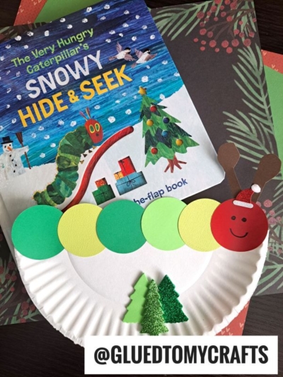 A copy of The Very Hungry Caterpillar's Snowy Hide & Seek book sits beside a Circle Punch Caterpillar Craft and a reindeer, both made from paper plates, decorated with colored circles, paper antlers, and small green Christmas trees.