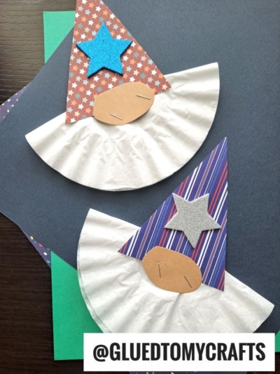 Two handmade coffee filter gnome crafts feature beards made from coffee filters, colorful paper hats, star decorations, and oval noses. They are displayed on dark and green paper backgrounds. @GLUEDTOMYCRAFTS is written at the bottom.