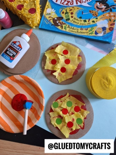 Two cardboard pizza craft circles are decorated with torn yellow paper and red and green dots. Glue, a paintbrush, yellow crepe paper, a painted plate, and a Pizza Ninja book are nearby. @GLUEDTOMYCRAFTS is shown at the bottom.