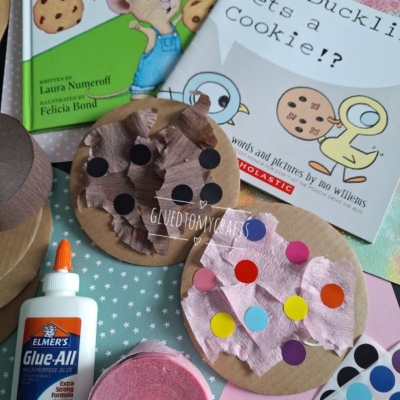Children’s books “If You Give a Mouse a Cookie” and “The Duckling Gets a Cookie!?” with craft supplies: paper cookies decorated with tissue and colored circles, Elmer’s glue, and pink crepe paper on a patterned surface.