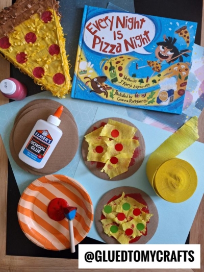 A children’s book titled Every Night Is Pizza Night with a cardboard pizza craft, plus other pizza-themed creations made from paper, paint, glue, and yellow streamers, displayed on a table with art supplies and an orange-striped plate.