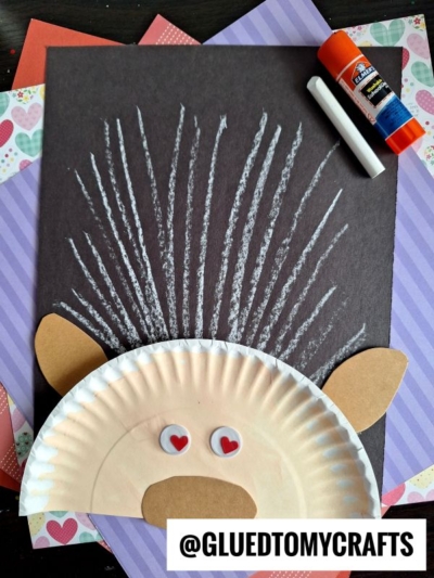 A paper plate porcupine craft—perfect for chalk art projects—with brown paper ears and nose, googly eyes with heart pupils, and white chalk quills on black paper. Glue stick, white chalk, and colorful paper are in the background.