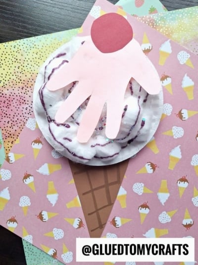 A cotton ball ice cream cone craft featuring a brown paper cone, a plate with fluffy cotton balls as ice cream, a pink handprint topping, and a red cherry sits on colorful ice cream-themed scrapbook paper. @GLUEDTOMYCRAFTS is labeled.