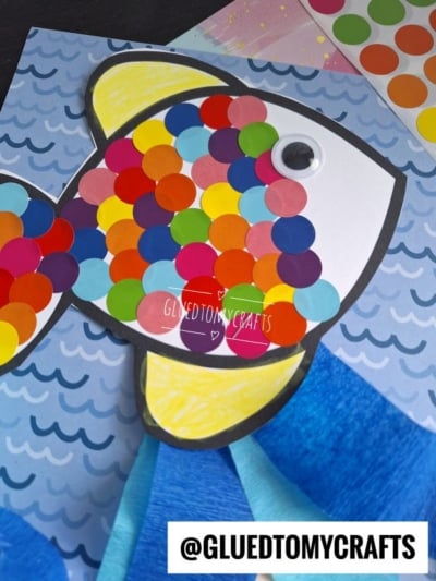 A colorful fish craft made from paper, decorated with bright round stickers as scales, a googly eye, and set on a blue wave-patterned background. The fish has yellow fins and a black outline.