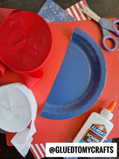 A blue paper plate cut in half, red and white crepe paper rolls, patterned paper, scissors, and a bottle of Elmer’s glue are arranged on a red polka dot surface. The Instagram handle @GLUEDTOMYCRAFTS appears at the bottom.