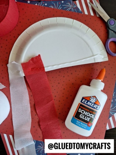 A white paper plate partially cut, red and white crepe paper strips, Elmer’s school glue, scissors, and craft materials arranged on a red and blue background.