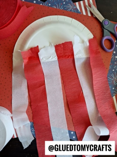 A paper plate with red and white crepe paper strips taped to it sits on red and blue star-patterned paper. Scissors and craft supplies are nearby. Text reads @GLUEDTOMYCRAFTS.