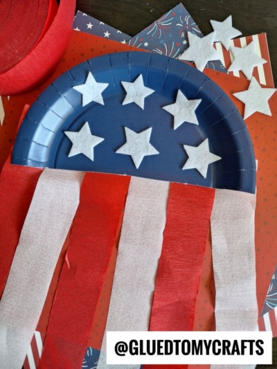 A blue paper plate decorated with white felt stars and red and white crepe paper streamers, resembling a patriotic American flag craft. Several stars and craft materials are scattered in the background.