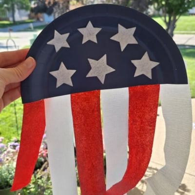 A hand holds a paper plate craft decorated like a U.S. flag, with blue and white stars on top and red and white streamers hanging down. Trees and grass are visible in the background. Text reads @GLUEDTOMYCRAFTS.