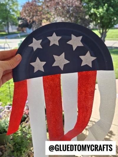 A hand holds a paper plate craft decorated like a U.S. flag, with blue and white stars on top and red and white streamers hanging down. Trees and grass are visible in the background. Text reads @GLUEDTOMYCRAFTS.