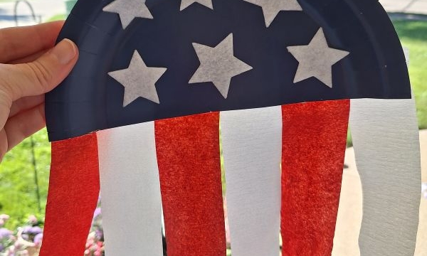 A hand holds a paper plate craft decorated like a U.S. flag, with blue and white stars on top and red and white streamers hanging down. Trees and grass are visible in the background. Text reads @GLUEDTOMYCRAFTS.